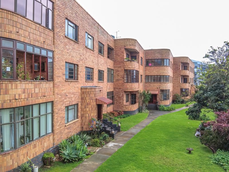 Exterior view of a classic brick apartment building featuring curved architectural details, steel-framed windows, and a well-maintained communal garden area.