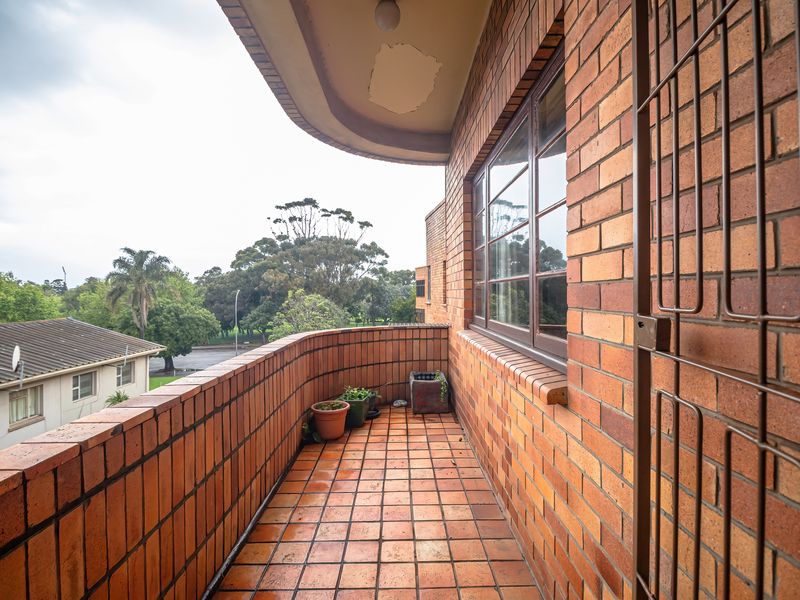 A private balcony featuring classic brickwork, tiled flooring, and a security gate, offering an outdoor space with views of the surrounding area.