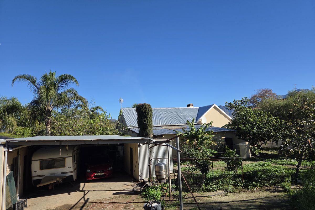 Rear view of the property featuring a corrugated metal roof, attached carport structure, and a large garden area with mature trees.
