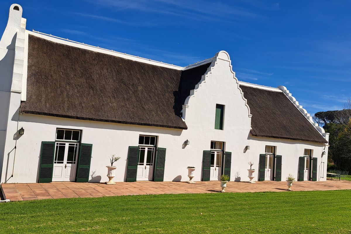 A well-maintained Cape Dutch style farmhouse featuring a traditional thatched roof, gabled architecture, and a spacious terracotta patio overlooking a manicured lawn.