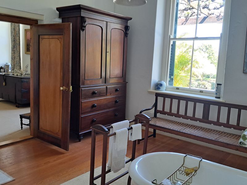 A spacious bathroom featuring classic timber flooring, a freestanding bathtub, and a large sash window overlooking the garden, with an adjoining room visible.