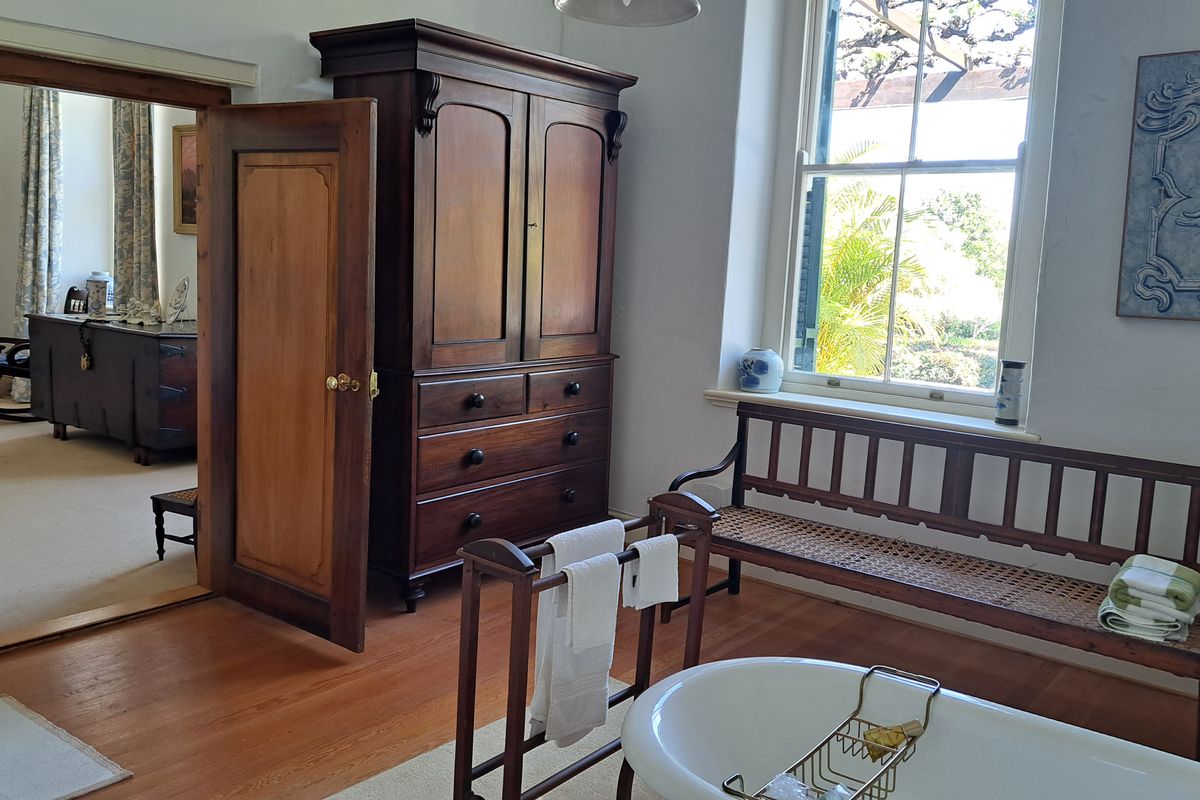 A spacious bathroom featuring classic timber flooring, a freestanding bathtub, and a large sash window overlooking the garden, with an adjoining room visible.