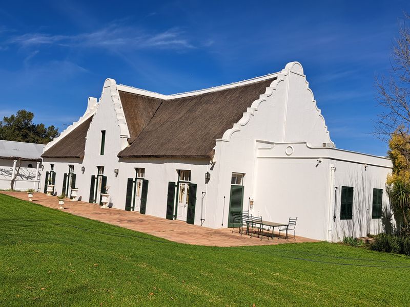 A well-maintained Cape Dutch style farmhouse featuring a traditional thatched roof, white gabled facade, and a spacious terracotta patio overlooking a green lawn.