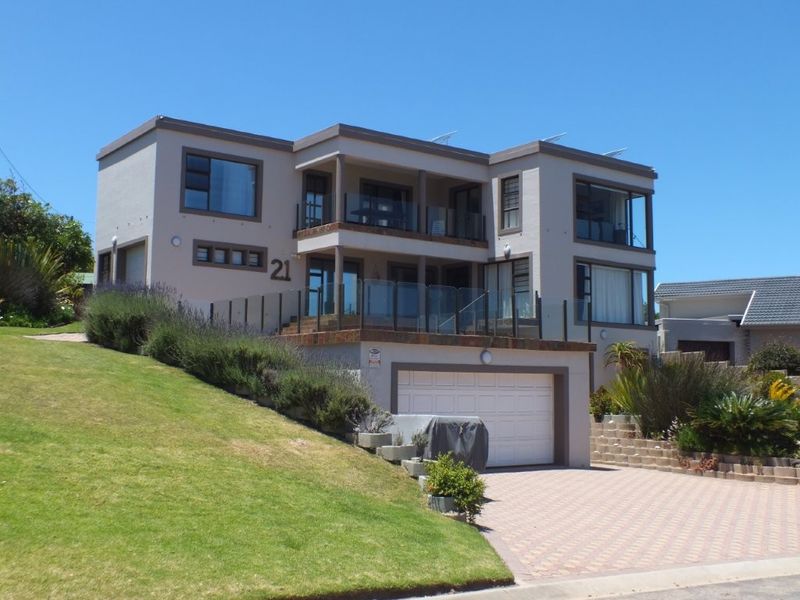 A multi-level residential property featuring a double garage, paved driveway, and tiered balconies with glass balustrades, set on a landscaped sloping plot.