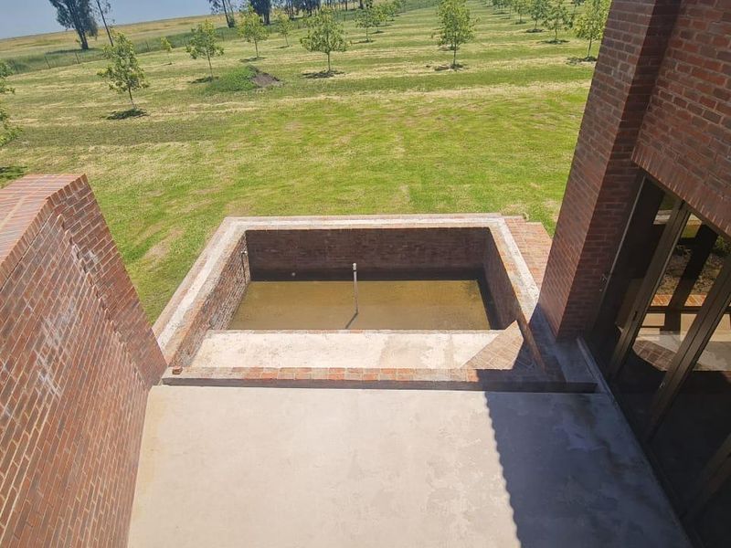 An elevated view of an unfinished brick swimming pool and concrete patio area, set against a large landscaped garden and open field.