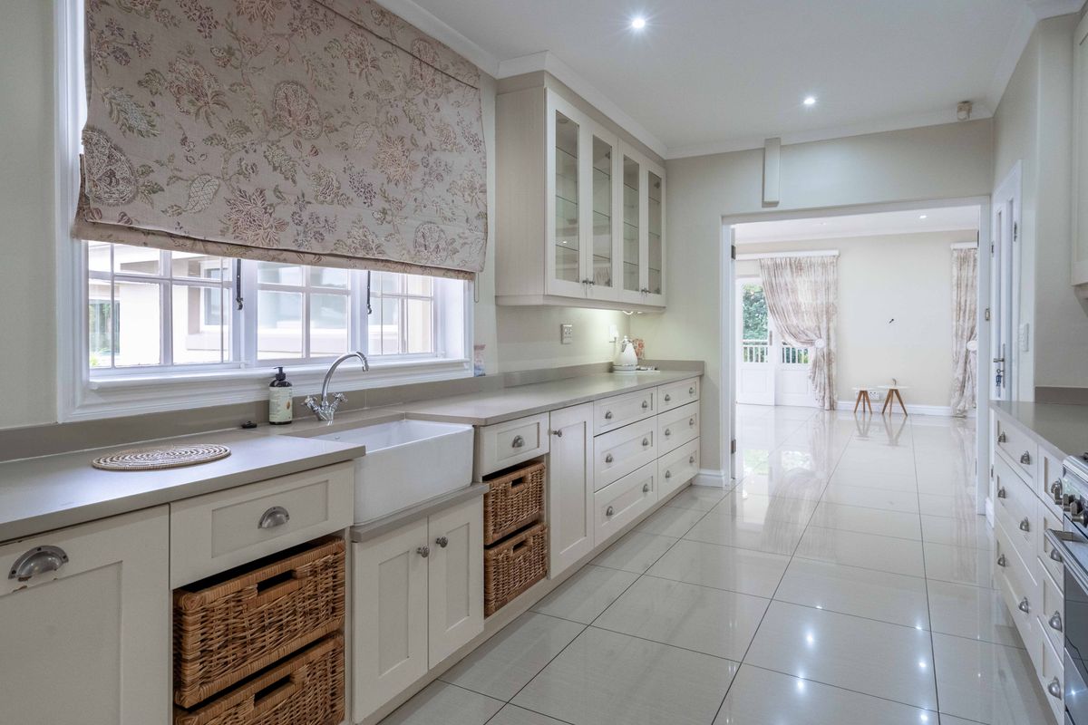 A bright, modern kitchen featuring shaker-style cabinetry, a classic farmhouse sink, stone countertops, and polished tile flooring with seamless flow to the adjacent living area.
