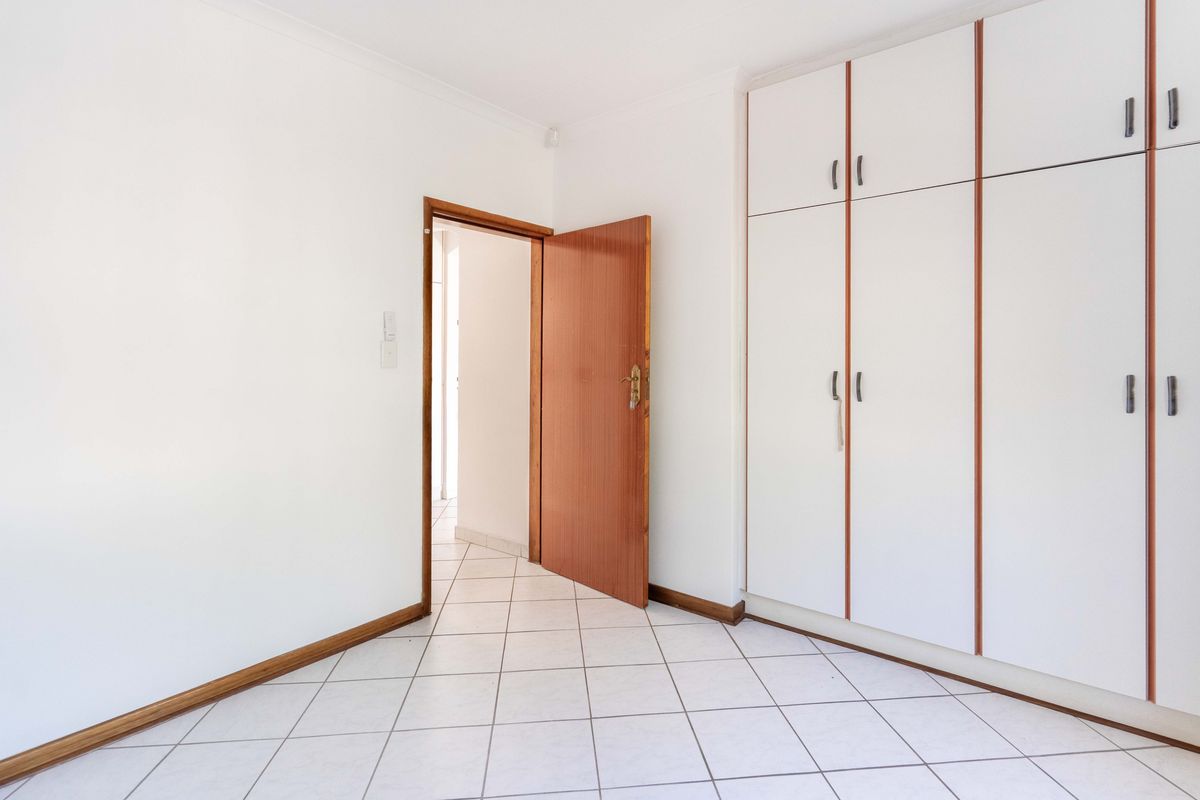 A bright, empty bedroom featuring white tiled flooring, built-in cupboards with white doors, and a wooden door leading to the hallway.