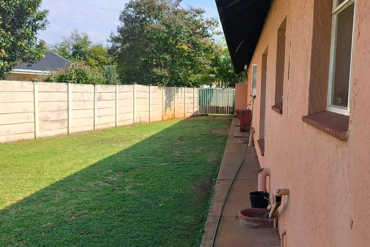 A rear exterior view of the property featuring a lawn, a precast concrete boundary wall, and a concrete pathway along the side of the house.