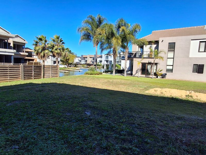 A rear view of a residential property featuring a grassy lawn, timber fencing, and mature palm trees with direct access to a waterfront area.