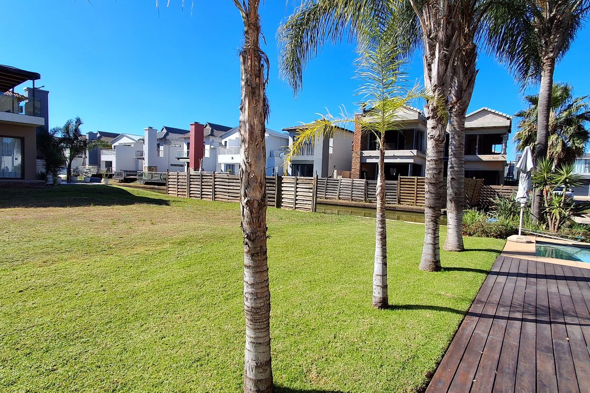 A well-maintained residential garden featuring a manicured lawn, mature palm trees, timber decking, and a swimming pool overlooking a canal.