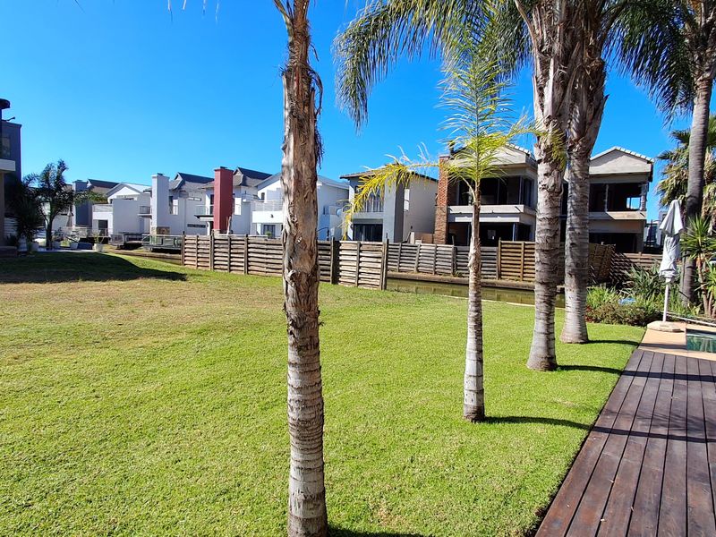 A well-maintained residential garden featuring a manicured lawn, mature palm trees, timber decking, and a swimming pool overlooking a canal.
