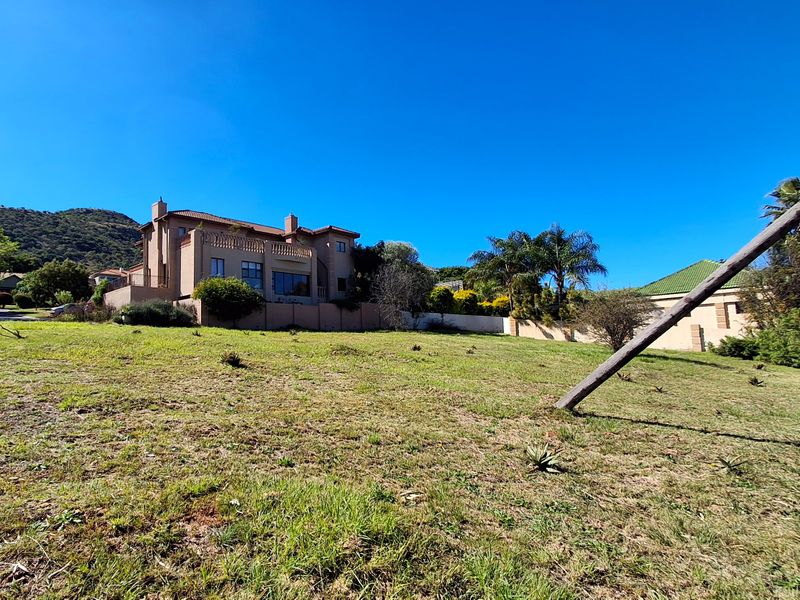 A view of a vacant residential plot with a gentle slope, featuring established boundary walls and surrounding residential properties in a scenic setting.