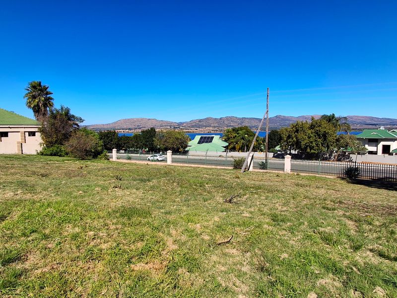 A wide-angle view of a vacant residential plot featuring established grass, perimeter fencing, and scenic mountain and water views in the background.