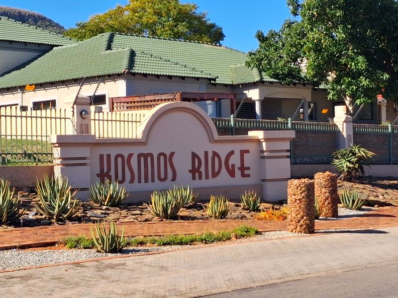 Entrance view of Kosmos Ridge estate featuring a prominent signage wall, security fencing, and well-maintained landscaped garden beds with stone gabion accents.