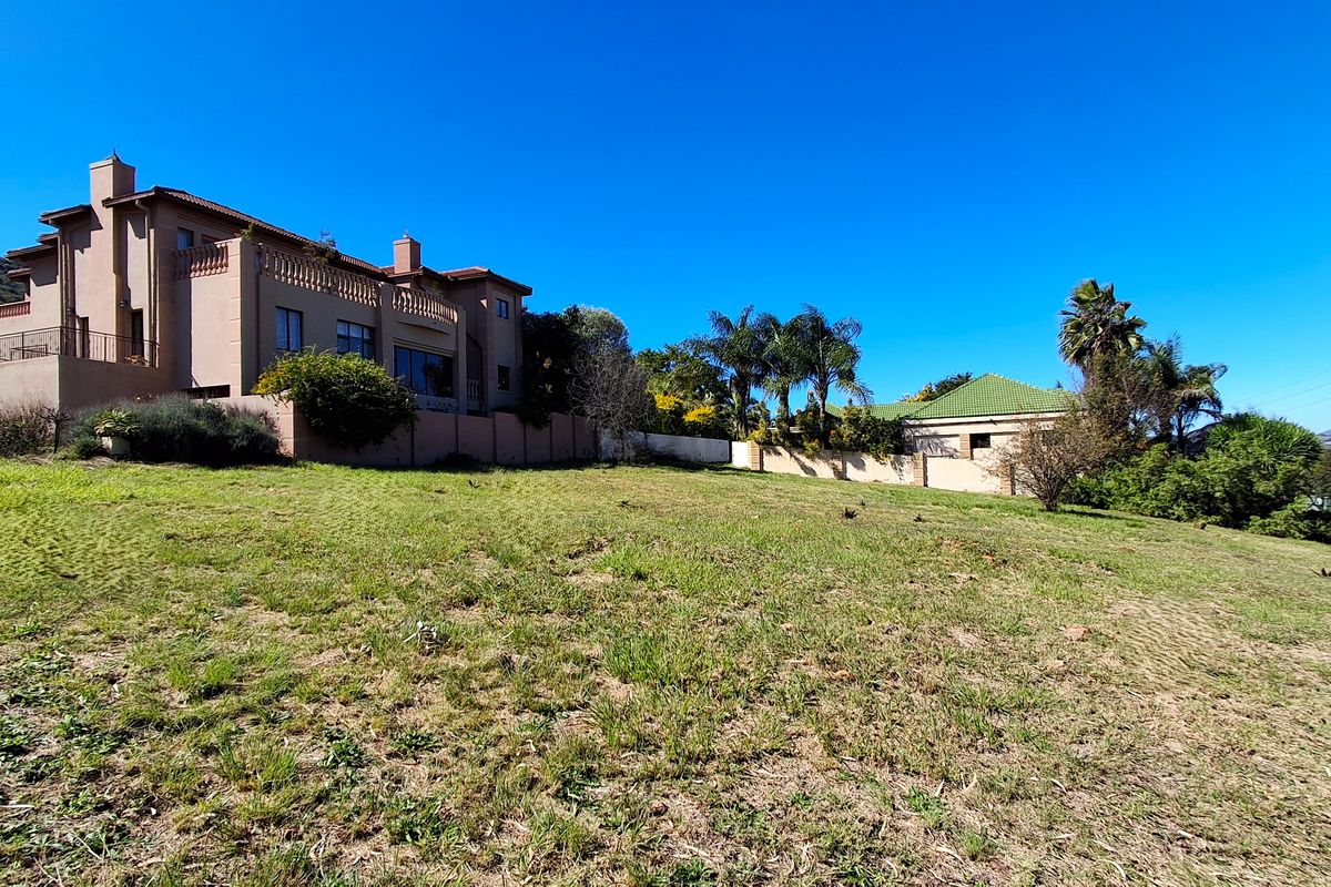 A view of a vacant residential plot featuring established garden landscaping and adjacent residential properties under a clear blue sky.