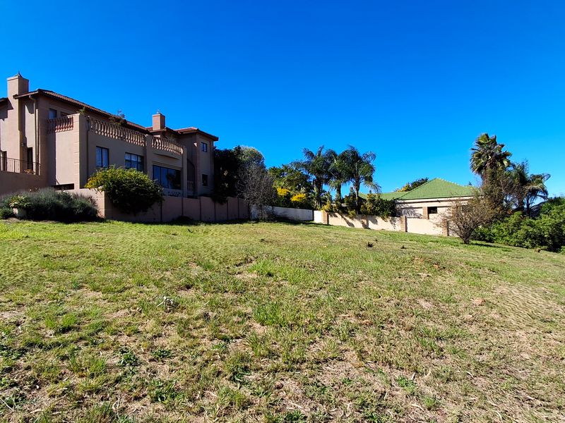 A view of a vacant residential plot featuring established garden landscaping and adjacent residential properties under a clear blue sky.