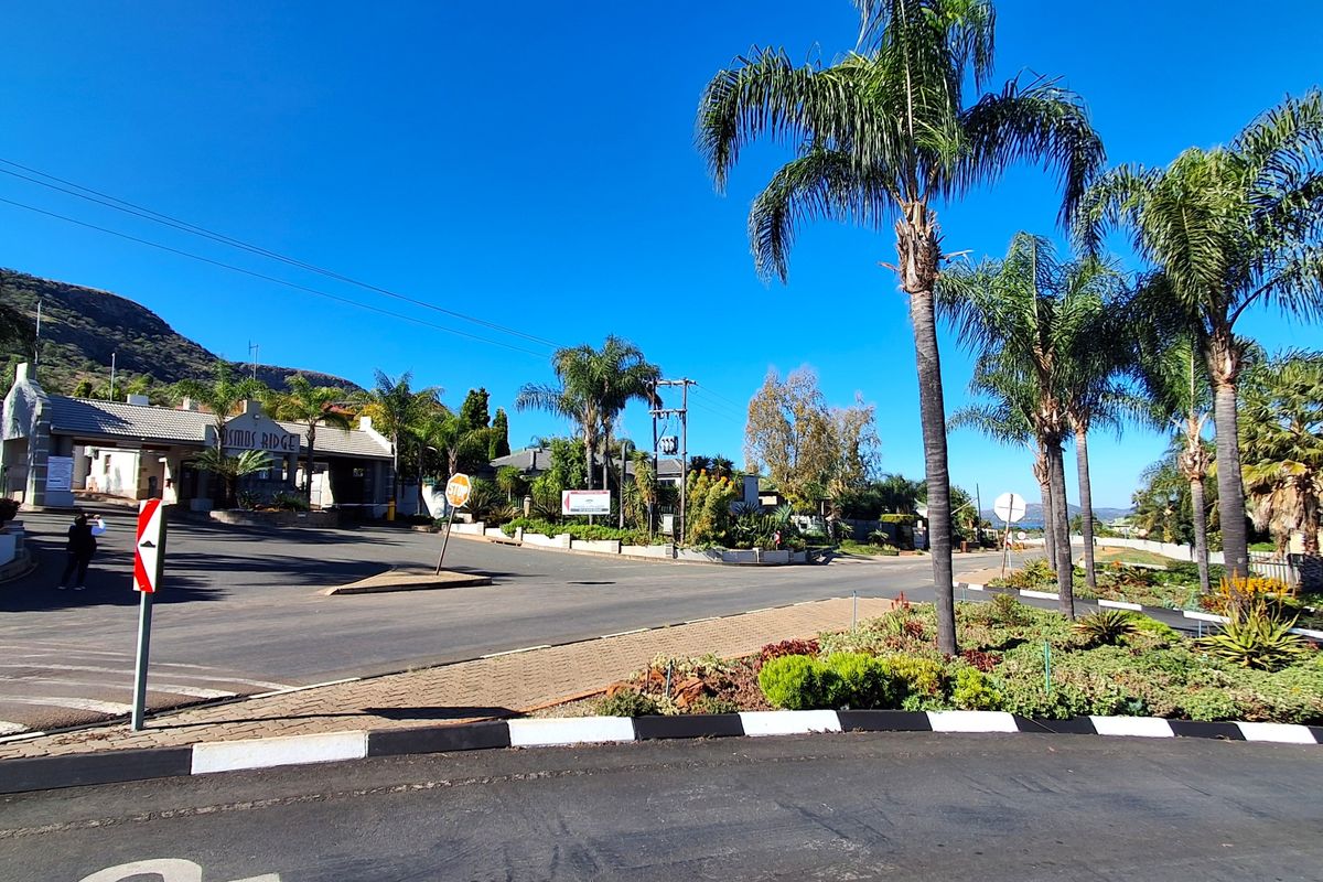 A view of the entrance to Cosmos Ridge, featuring a paved road, landscaped verge with palm trees, and a security gatehouse.
