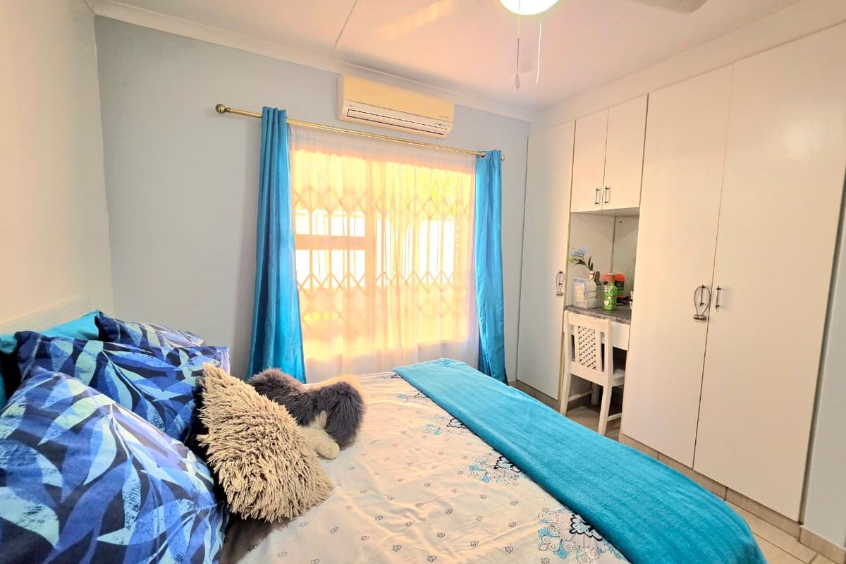 A functional bedroom featuring built-in cupboards, a ceiling fan, and a wall-mounted air conditioning unit, with tiled flooring and security window treatments.