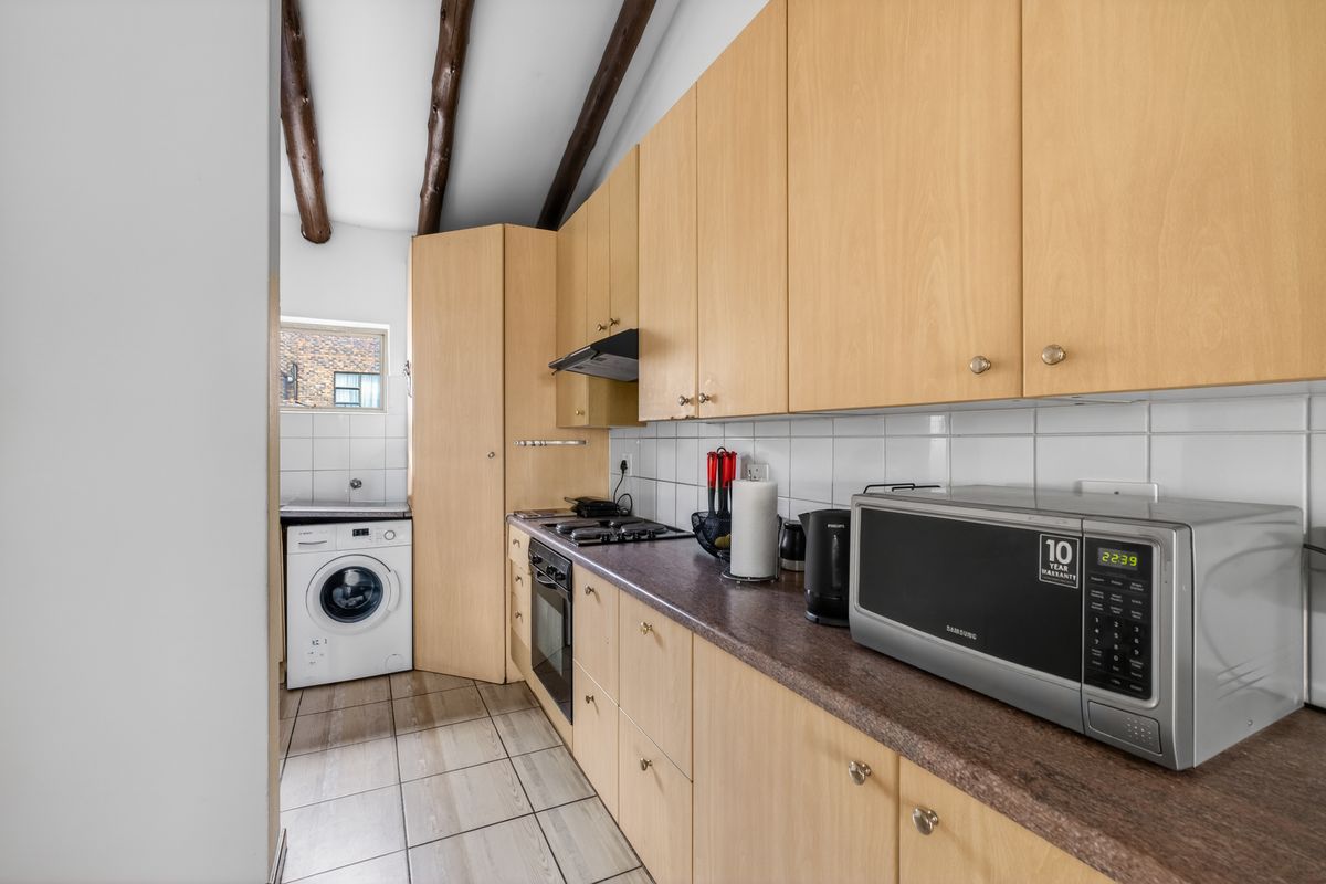A functional kitchen featuring light wood cabinetry, tiled backsplash, built-in oven and hob, exposed timber ceiling beams, and a dedicated laundry area.
