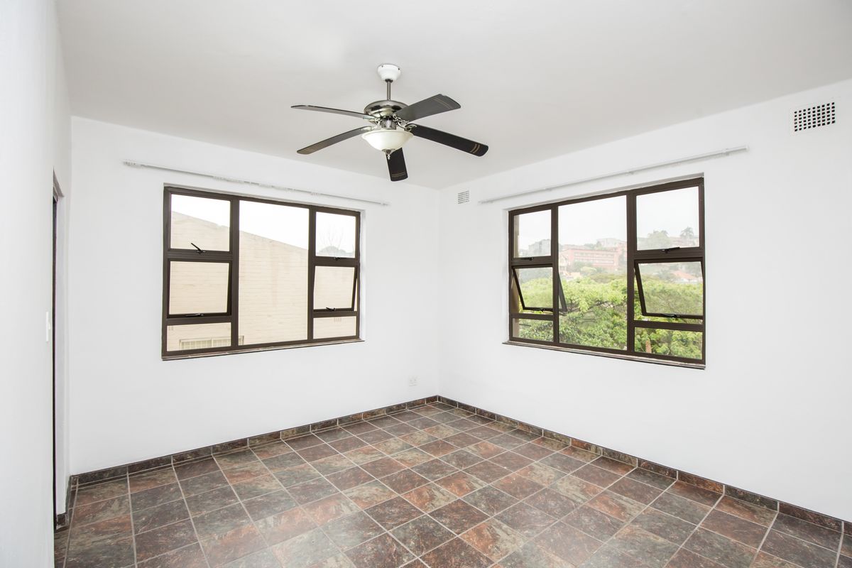 A bright, empty bedroom featuring tiled flooring, large windows with dark frames, and a ceiling fan.