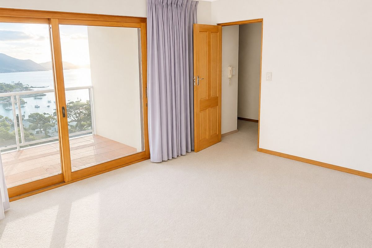 A bright, empty bedroom featuring neutral carpeting, timber door frames, and a large sliding glass door providing direct access to a balcony with water views.