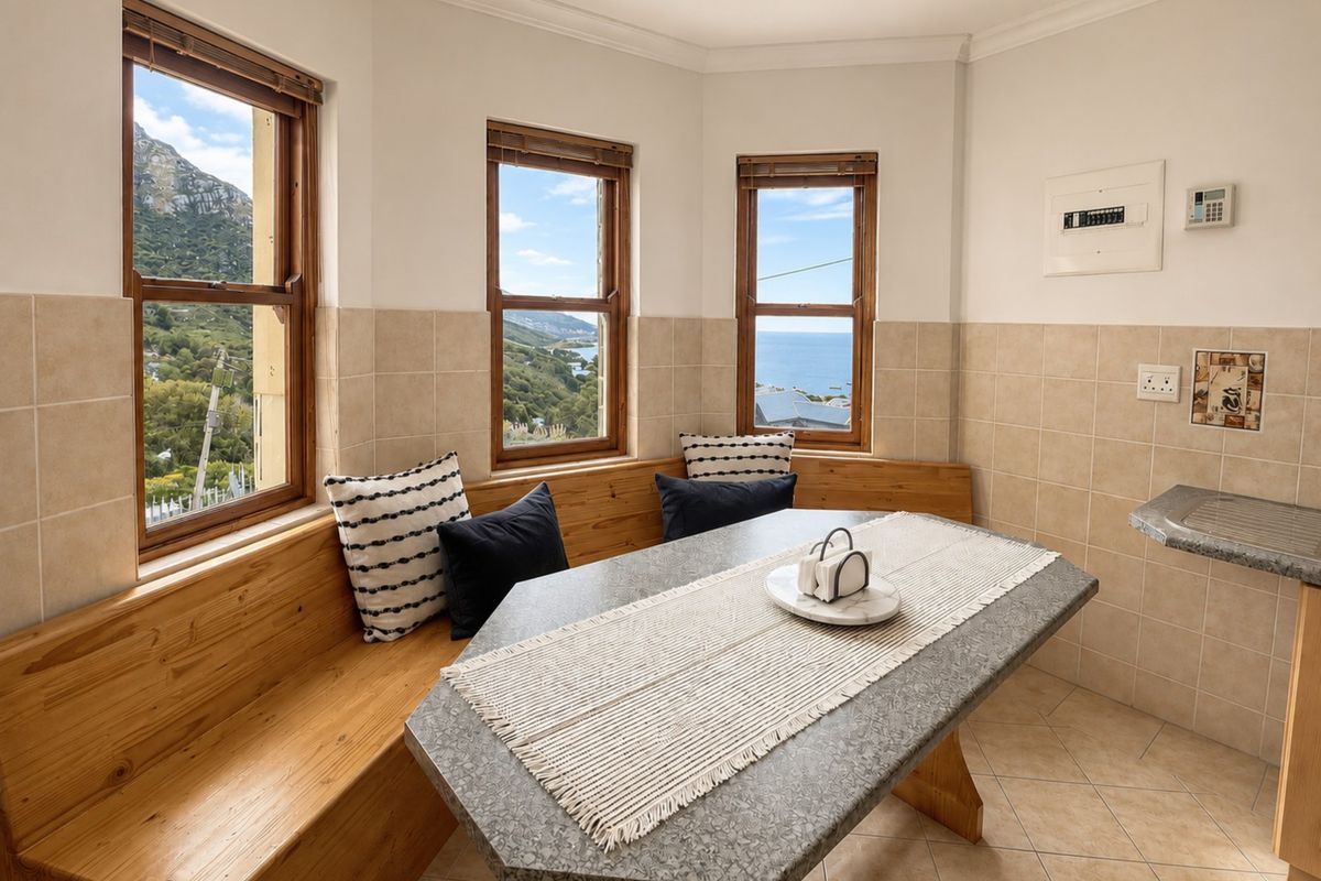 A kitchen breakfast nook featuring built-in timber bench seating, a stone-look dining table, and large windows offering scenic coastal and mountain views.