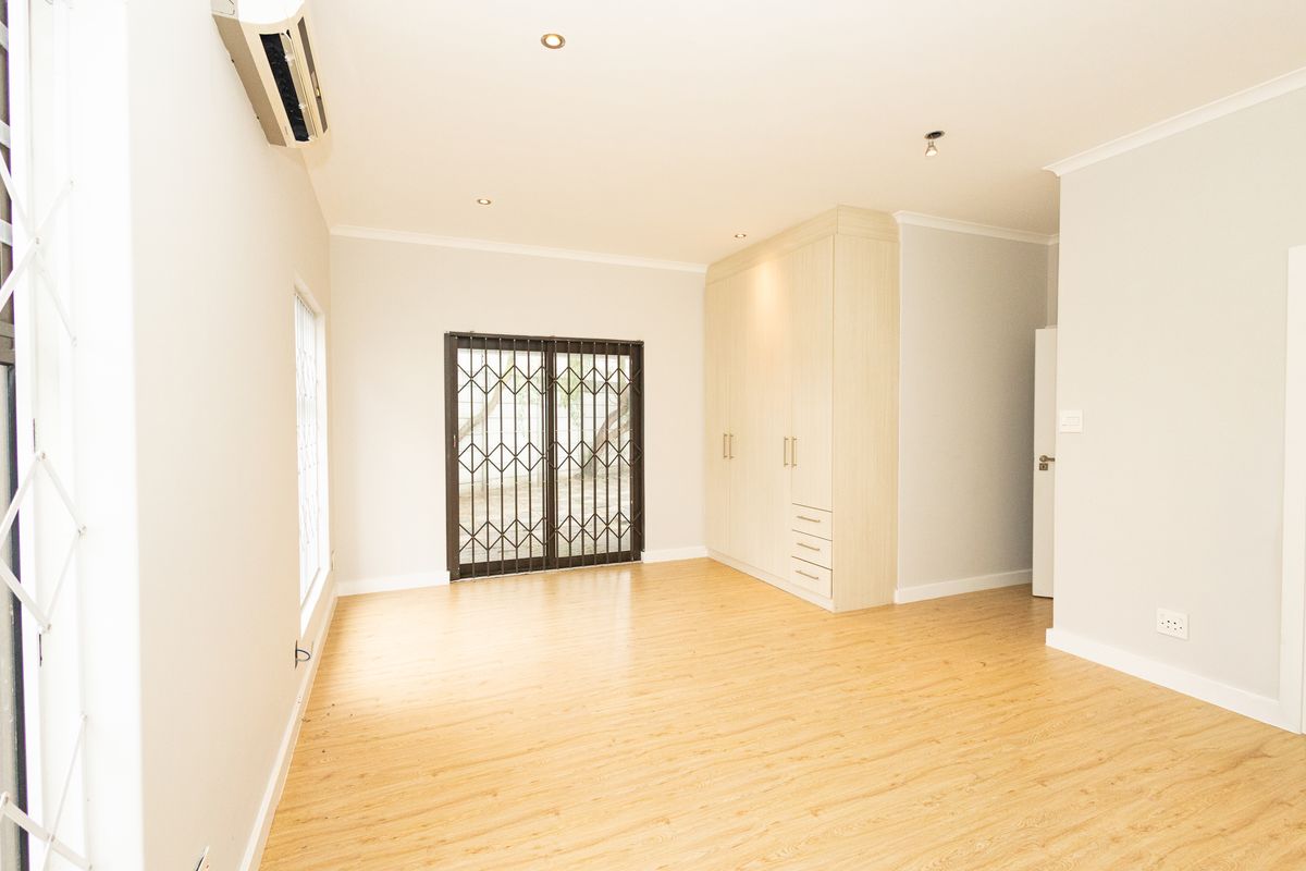 A bright, empty bedroom featuring light-toned laminate flooring, built-in cupboards, and an air conditioning unit, with a security gate leading to the exterior.