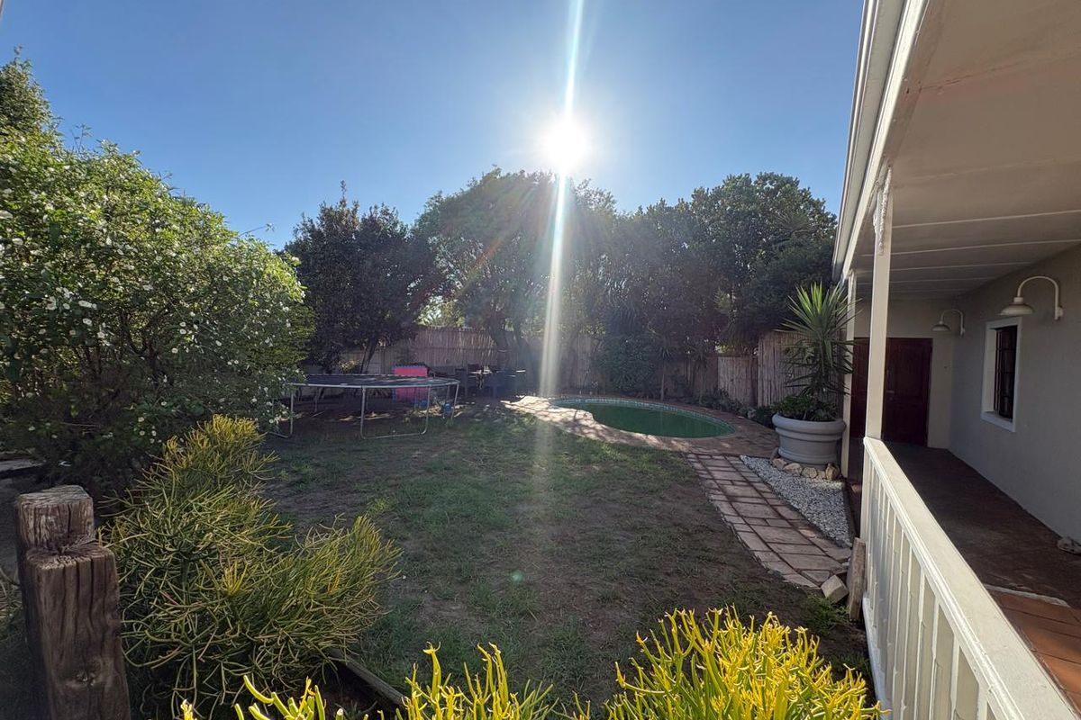 A rear garden featuring a swimming pool, paved walkway, and a covered patio area, set within a private, tree-lined backyard.