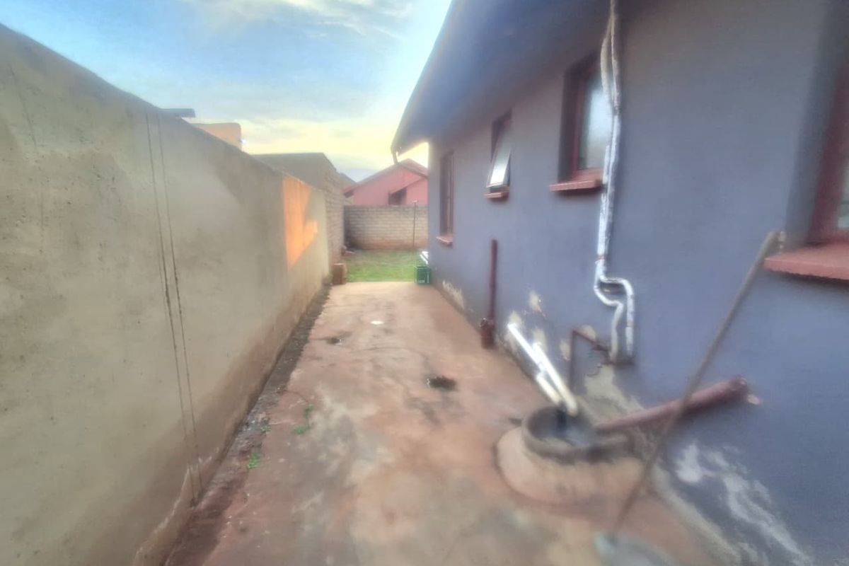 A narrow rear exterior pathway featuring a concrete perimeter wall, external plumbing fixtures, and a concrete walkway requiring maintenance and surface repairs.