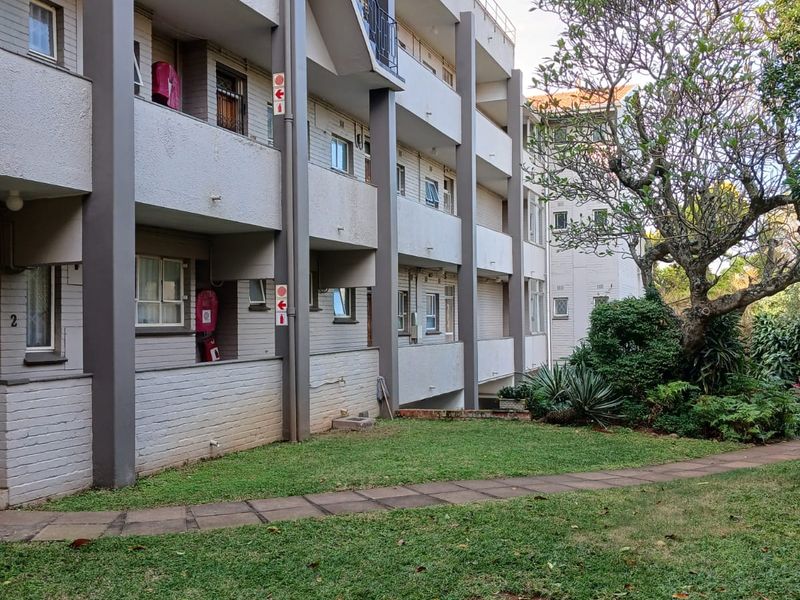 Exterior view of an apartment building featuring concrete balconies, an external stairwell, and a well-maintained garden area with a paved walkway.