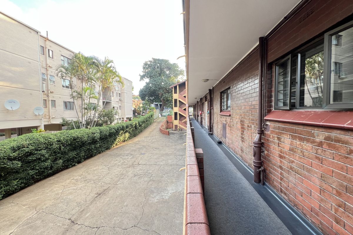 Exterior view of an apartment building walkway featuring brick facade, concrete flooring, and metal safety railings along the building perimeter.