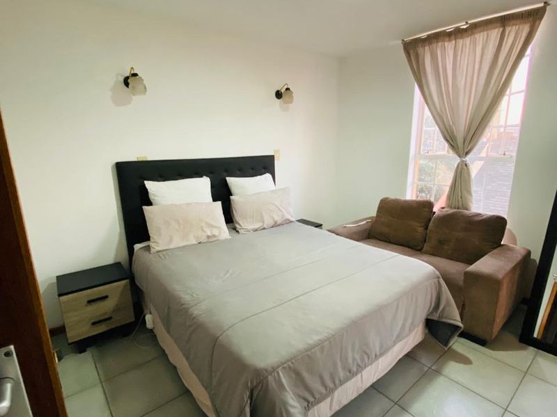 A bedroom featuring tiled flooring, wall-mounted lighting, and a window with curtains, providing a functional and well-lit sleeping space.