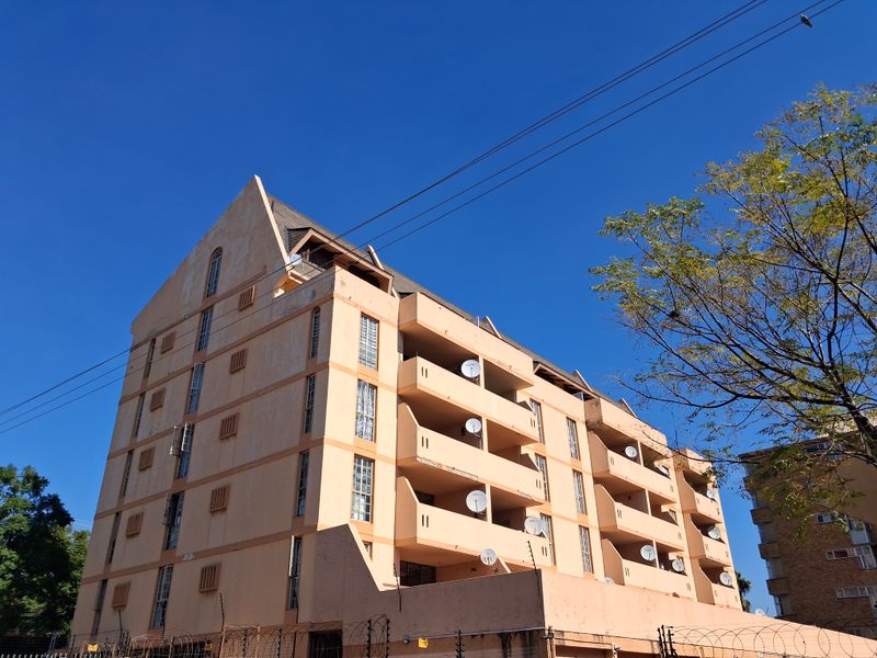 Exterior view of a multi-story apartment building featuring stacked balconies, a pitched roof design, and perimeter security fencing.