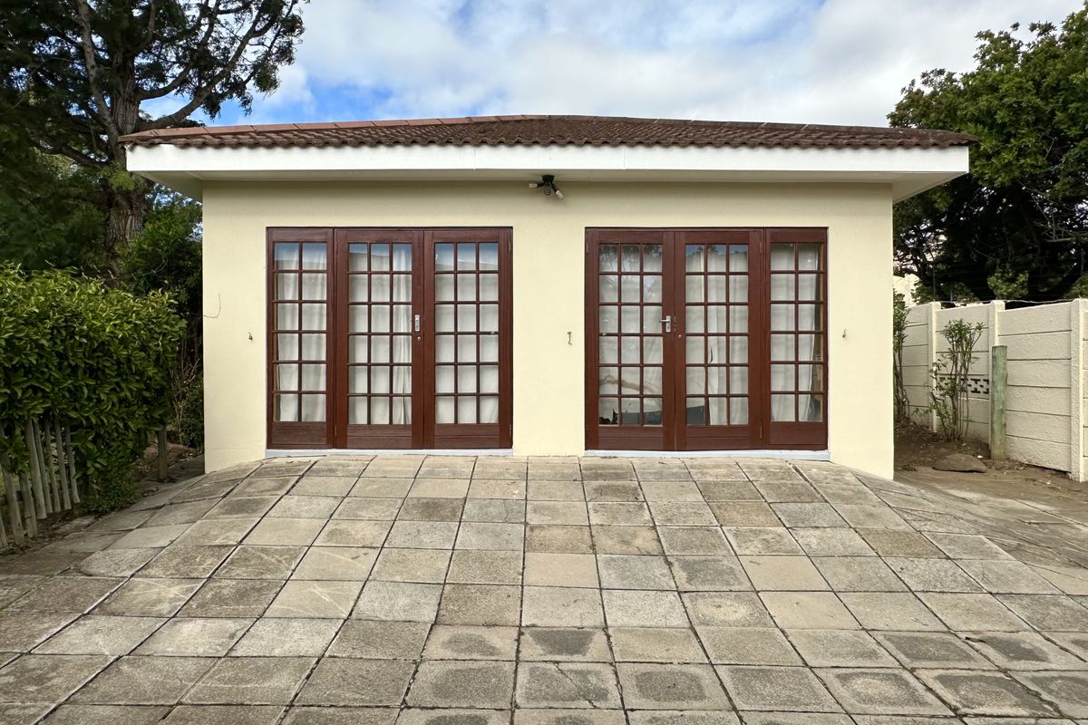 Rear view of a detached outbuilding featuring a tiled roof and two sets of wooden french doors opening onto a paved patio area.