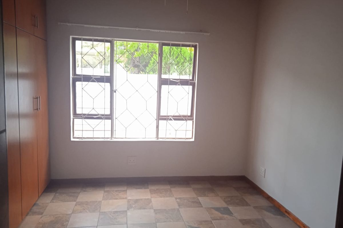 A clean, empty bedroom featuring built-in cupboards, tiled flooring, and a window fitted with security bars.