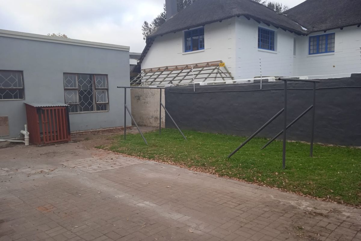 Rear exterior view of the property featuring a paved driveway, lawn area, and a building with a thatched roof and security features.