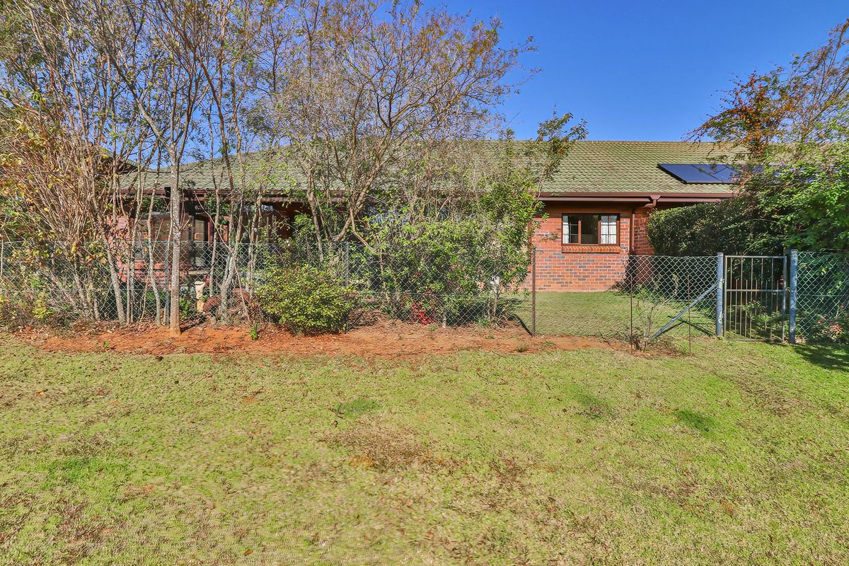 Rear view of a brick retirement village unit featuring a tiled roof with solar panels, enclosed by chain link fencing and mature garden landscaping.