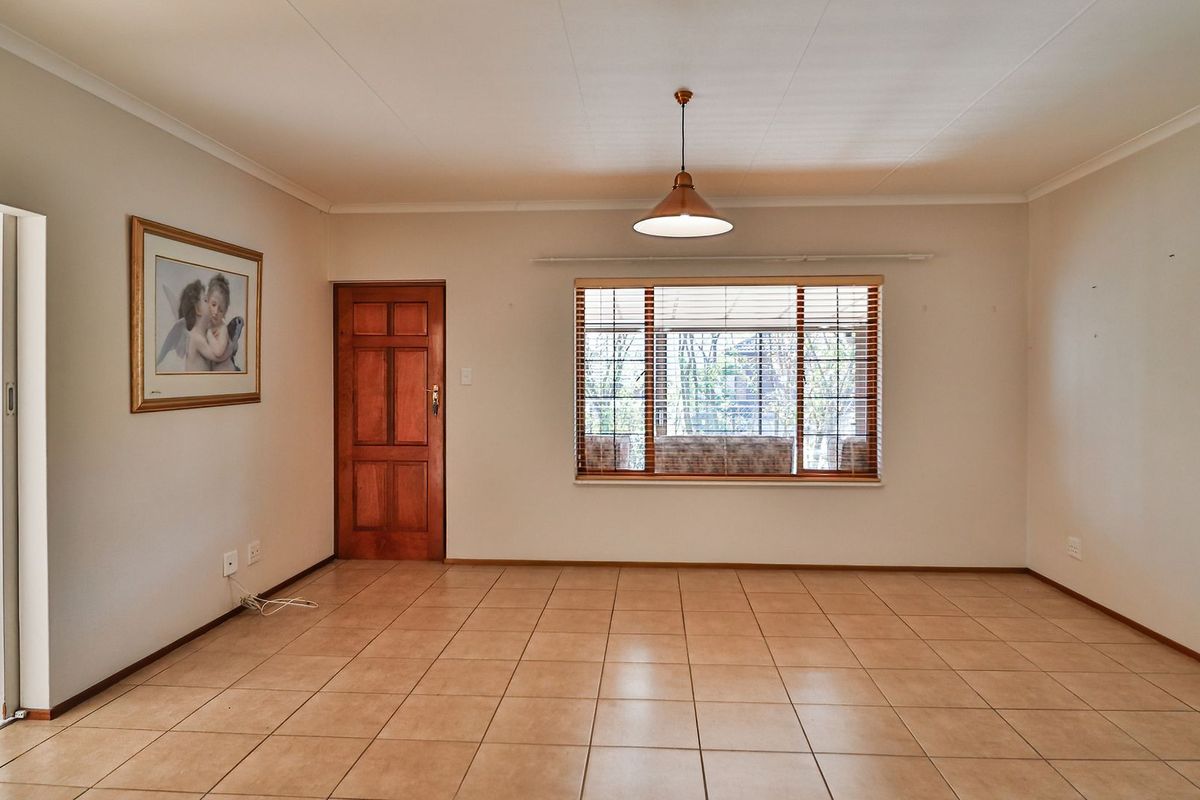 A spacious, empty living room featuring neutral tiled flooring, a central pendant light, and a wooden door leading to an adjacent space.