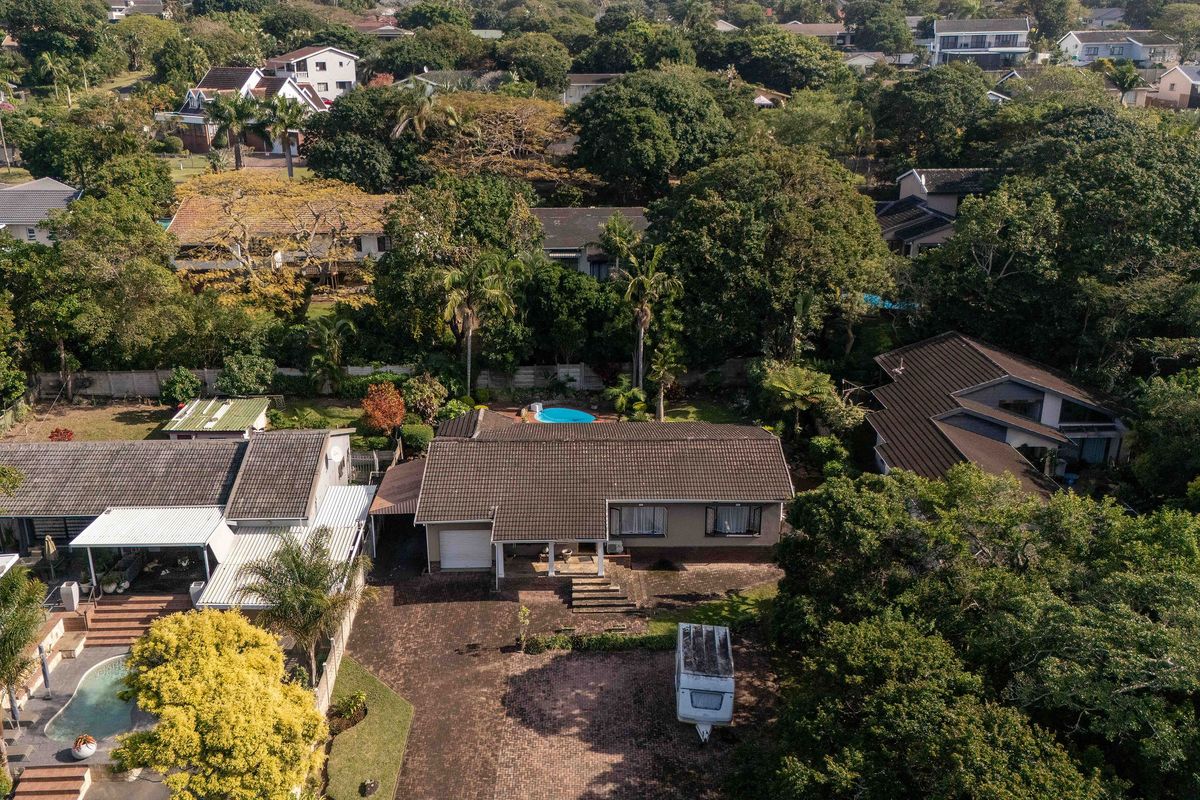 Aerial view of a residential property featuring a tiled roof, paved driveway, garage, and a private swimming pool set within a lush, tree-lined neighborhood.