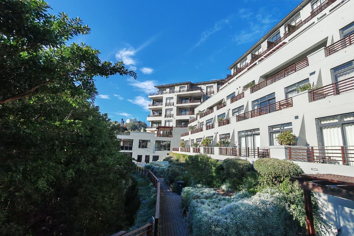Exterior view of a multi-story apartment complex featuring landscaped gardens, paved walkways, and private balconies with metal railings.