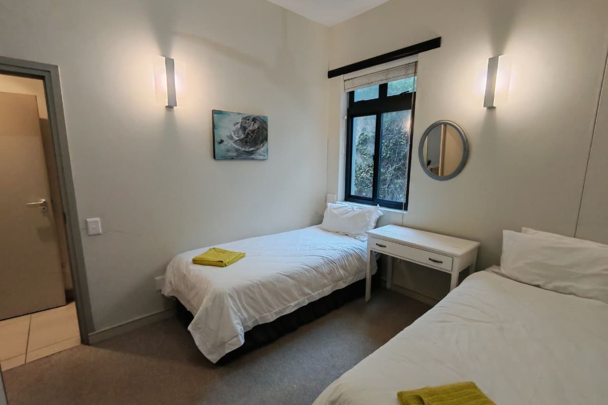 A functional bedroom featuring neutral wall tones, carpeted flooring, wall-mounted lighting fixtures, and a window with a view of exterior greenery.