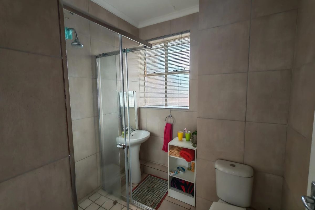 A functional bathroom featuring a glass-enclosed shower, pedestal basin, and toilet with neutral wall tiling and a window for natural light.