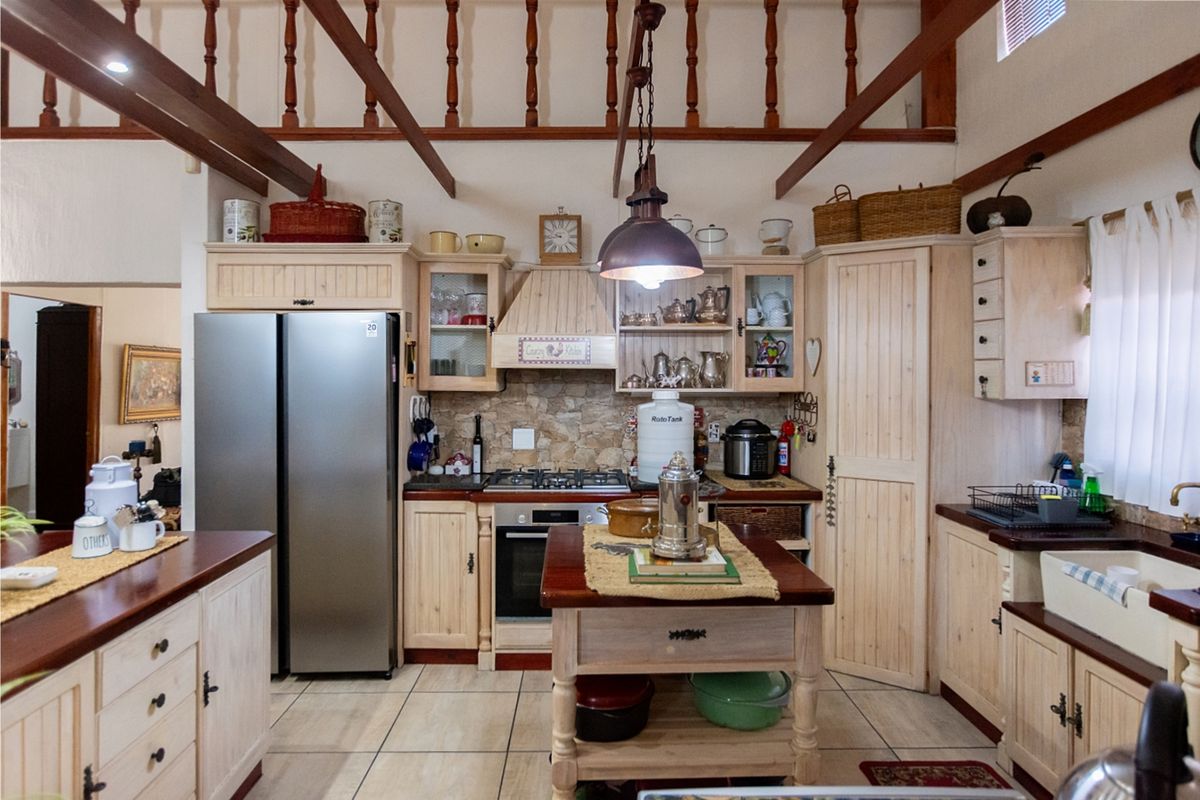 A rustic-style kitchen featuring light timber cabinetry, stone backsplash, farmhouse sink, and exposed timber ceiling beams with a central island.