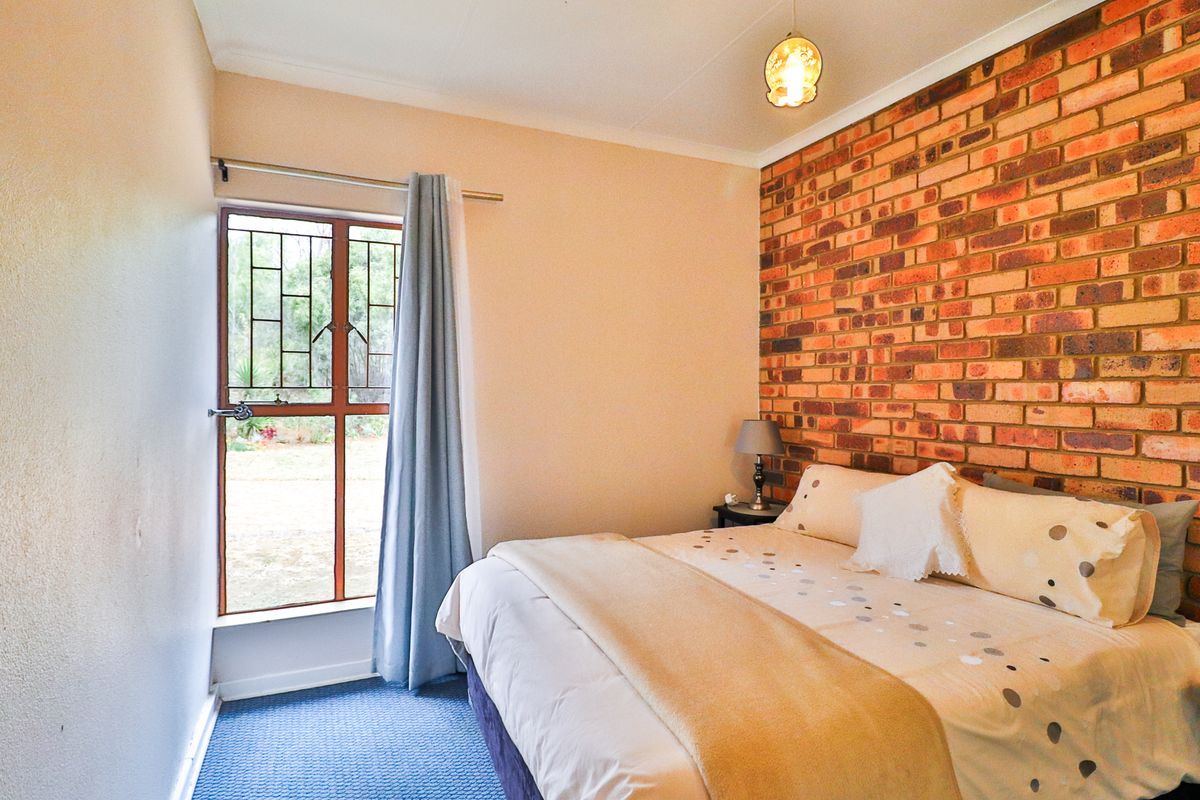 A bedroom featuring an exposed brick accent wall, carpeted flooring, and a window with security bars overlooking the garden.