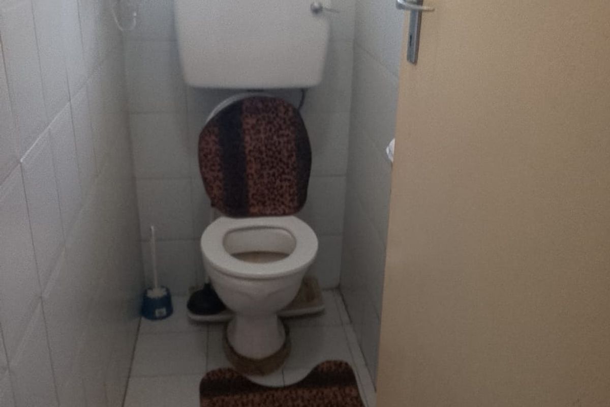 A small, functional bathroom featuring white ceramic wall and floor tiles, a standard toilet with cistern, and a window with security bars.