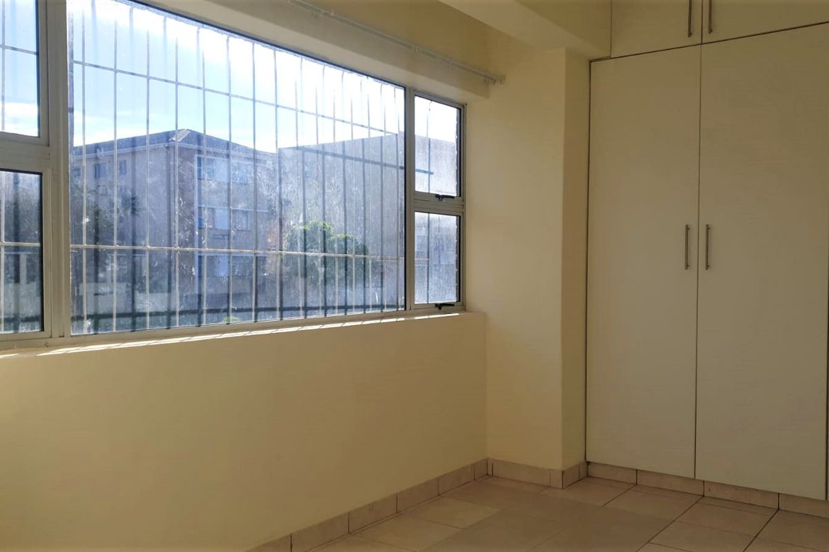 A bedroom featuring built-in cupboards, tiled flooring, and large windows with security bars, offering natural light and a view of the surrounding complex.