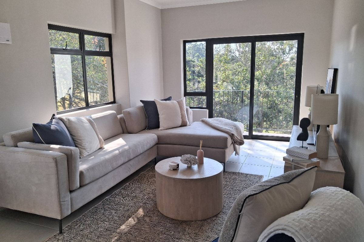 A bright and well-maintained living room featuring tiled flooring, large windows, and a sliding glass door leading to a balcony with leafy views.