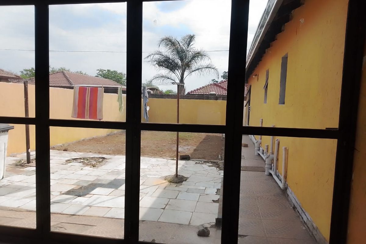 View of a rear courtyard featuring paved areas, exposed exterior plumbing, and a perimeter wall, viewed through a large window frame.