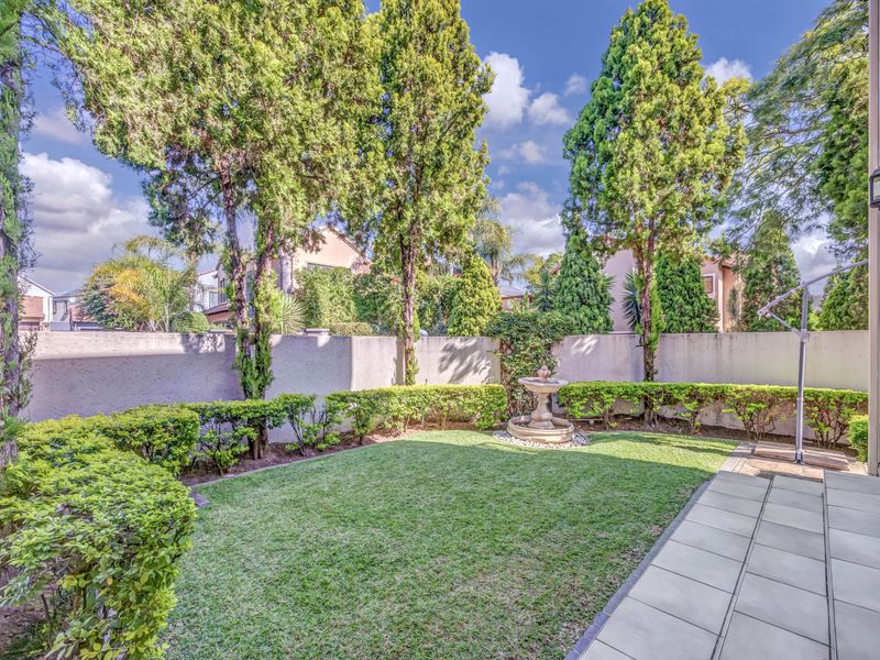 A well-maintained private garden featuring a manicured lawn, mature trees, perimeter walling, and a central stone fountain, adjacent to a paved patio area.