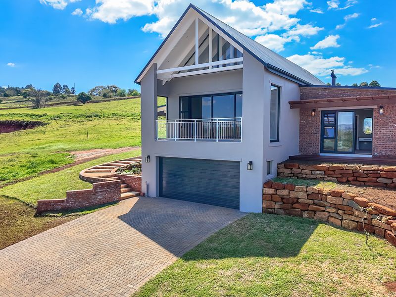 Modern two-story home featuring a paved driveway, double garage, upper-level balcony, and a combination of rendered and exposed brick finishes set in a rural landscape.